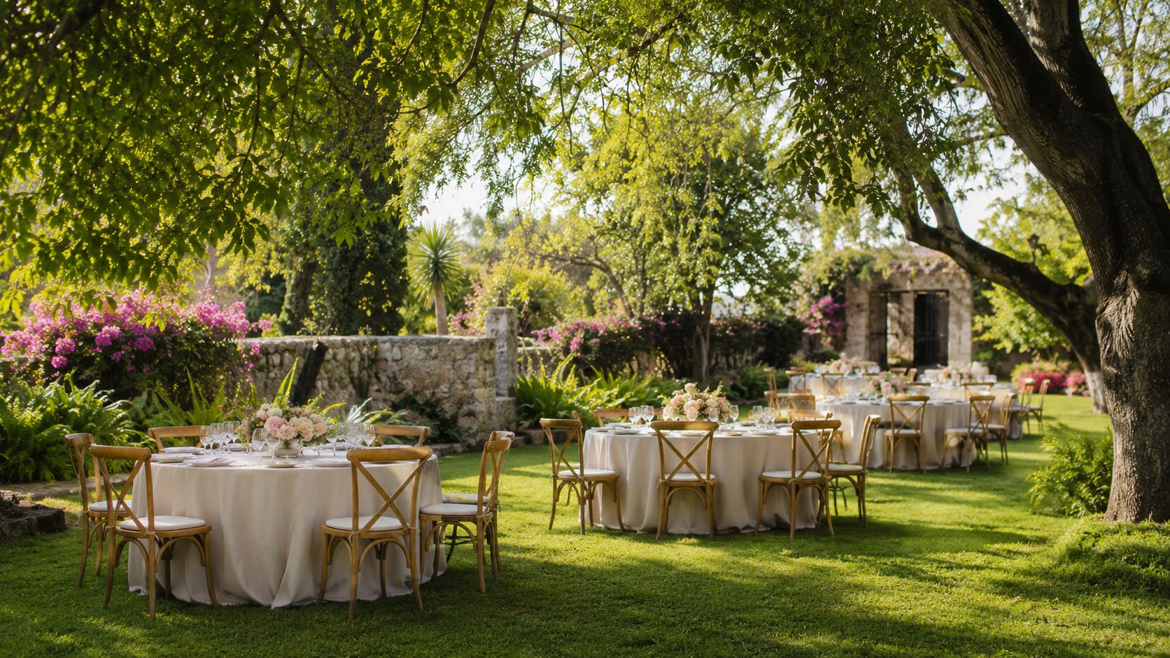 Mesas redondas decoradas para un evento al aire libre en un hermoso jardín con árboles frondosos.